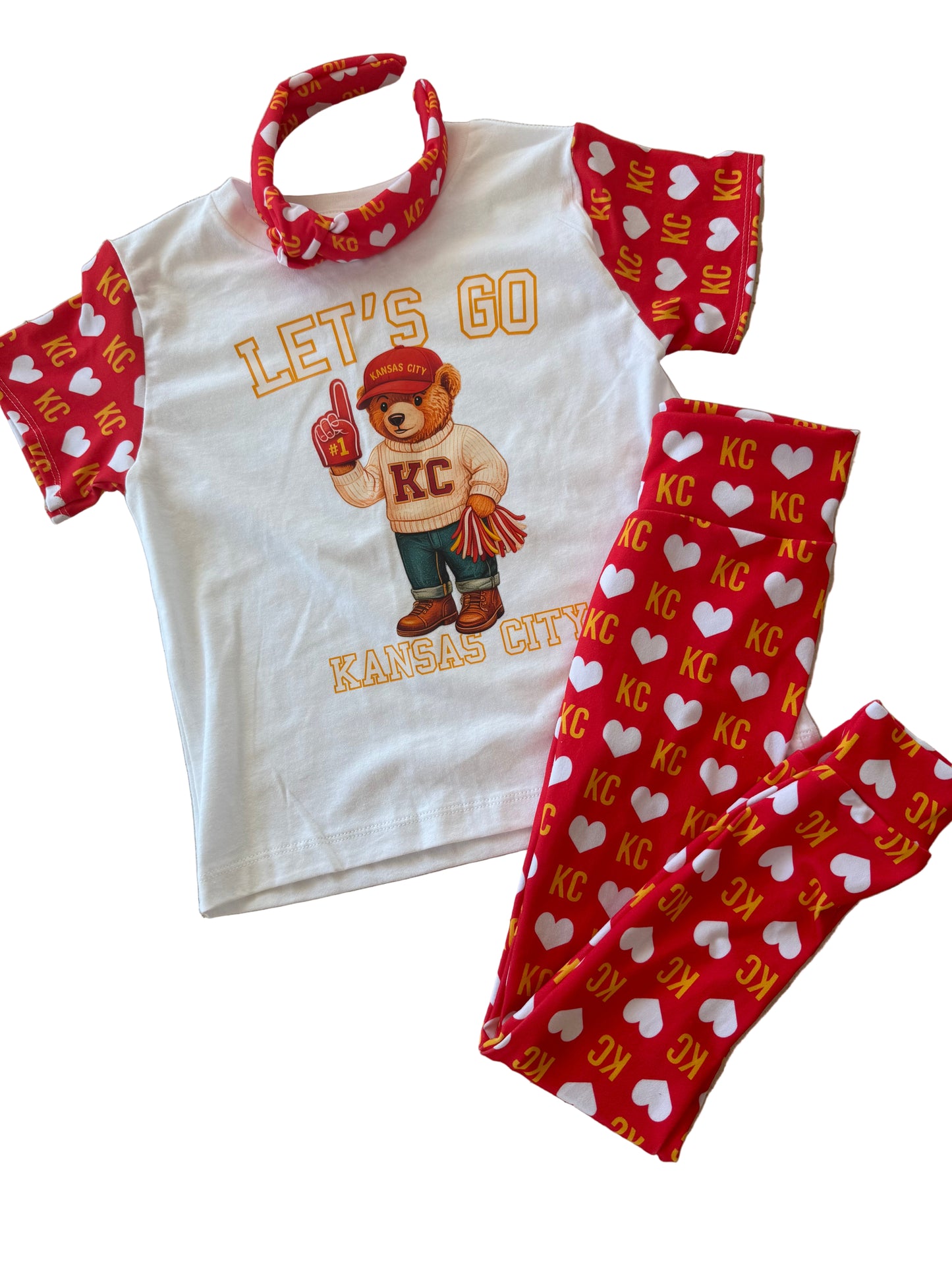 White t-shirt with bear graphic and 'Let's Go Kansas City' text, red pants with white heart patterns, and red headband