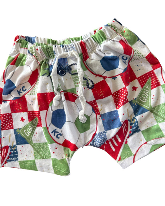A pair of children's shorts featuring a colorful patchwork pattern with soccer balls, green checkered flags, and red, white, and blue squares, complete with a white drawstring.