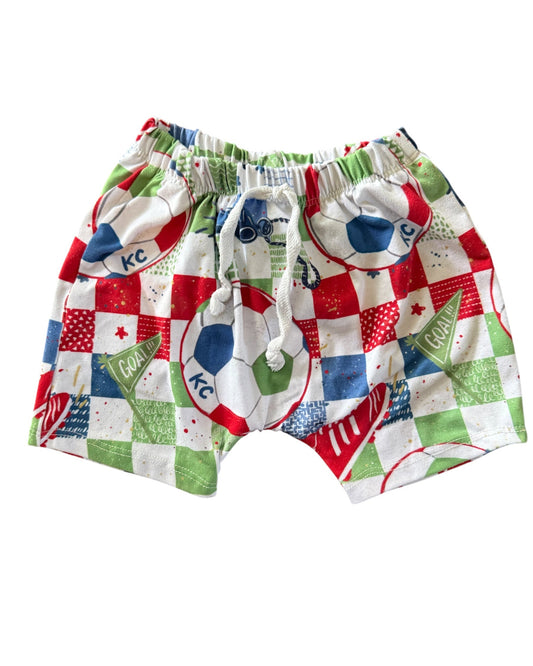 A pair of children's shorts featuring a colorful patchwork print with soccer balls, checkered squares, and green grass motifs in red, white, blue, and green. The shorts have an elastic waistband with a white drawstring.