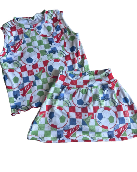 A sleeveless white top and matching skirt set with a colorful soccer-themed patchwork print, laid flat on a brown carpet.