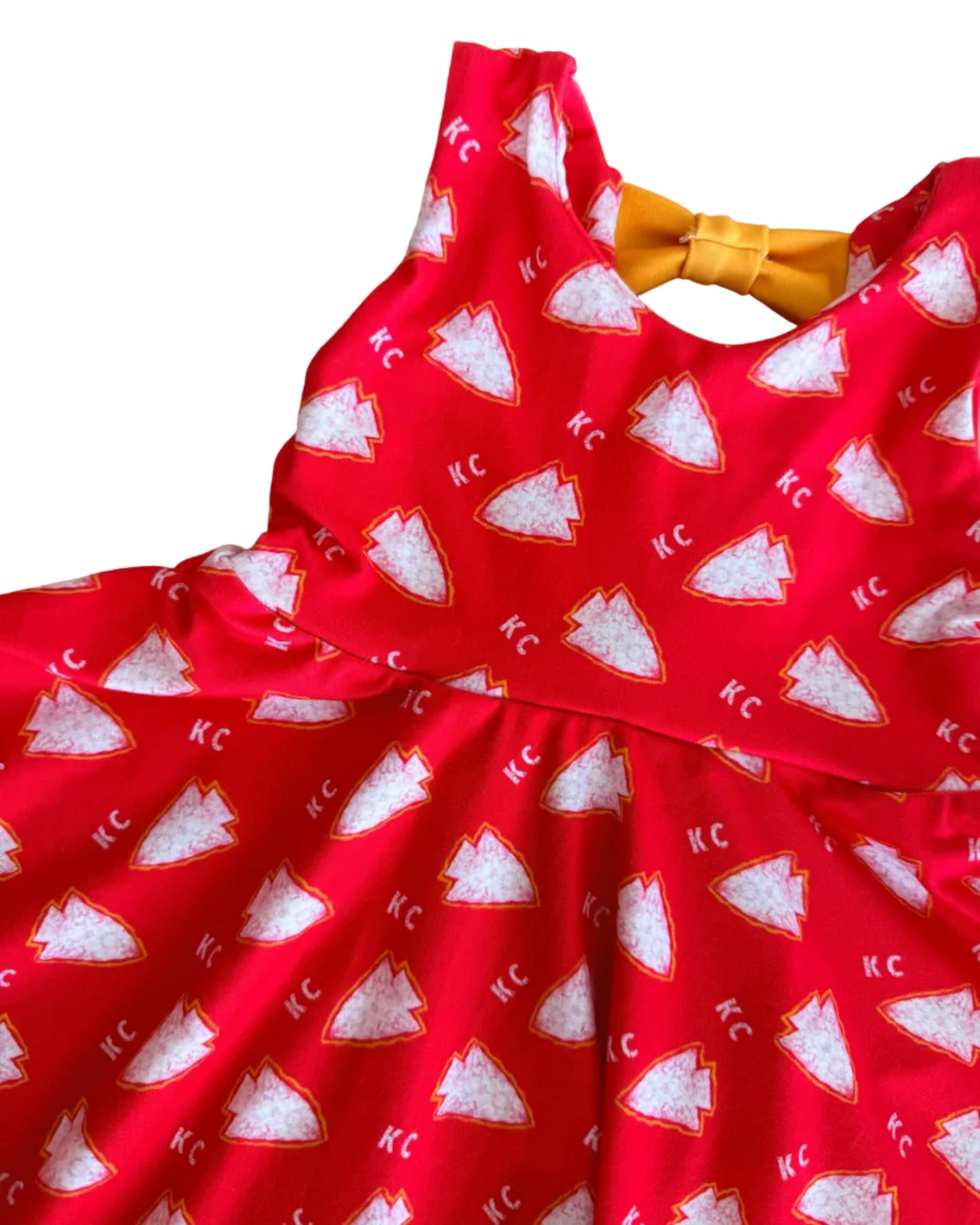 A red sleeveless dress with a repeating white  arrowhead pattern and scattered 'KC' text, featuring a yellow bow at the neckline, laid flat on a white floor.