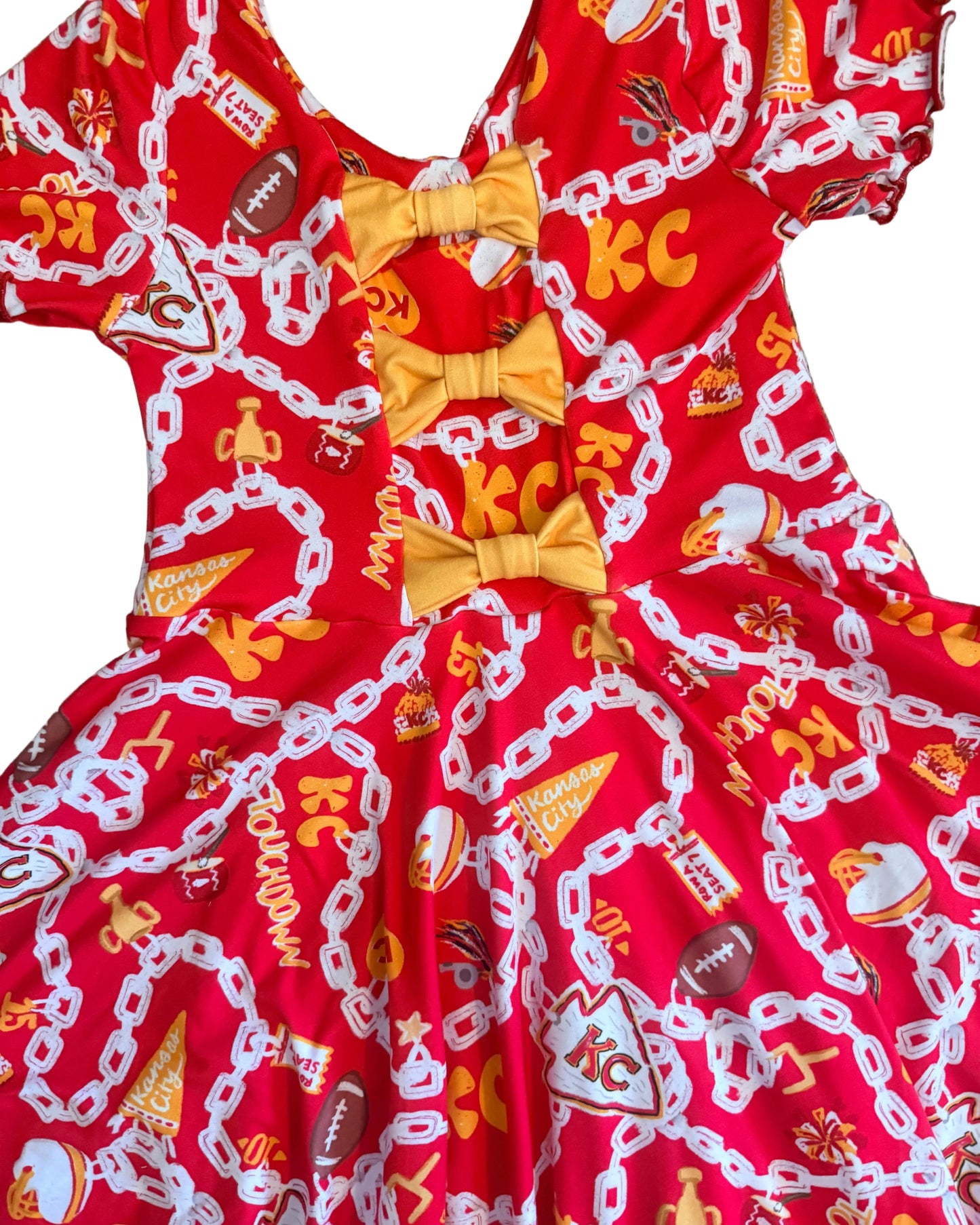 A red fabric dress with a football-themed print featuring white chain graphics, orange and yellow details, and three yellow bow accents at the center, laid flat on white carpet.