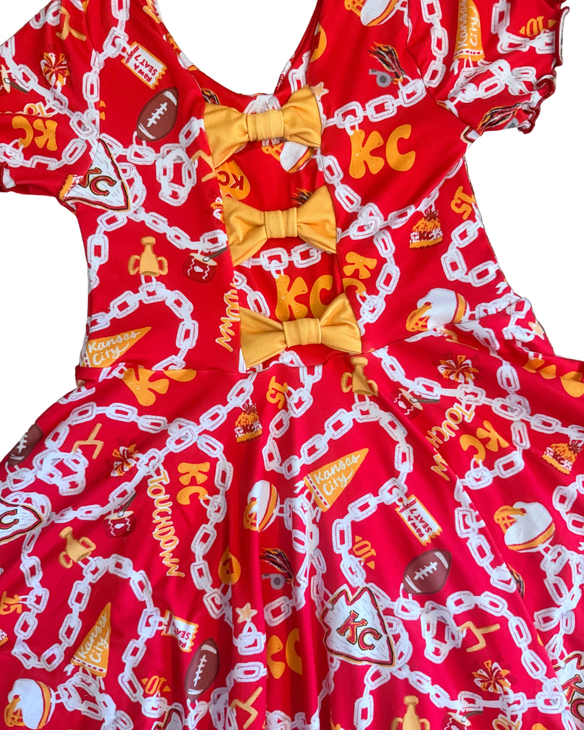 A red fabric dress with a football-themed print featuring white chain graphics, orange and yellow details, and three yellow bow accents at the center, laid flat on white carpet.