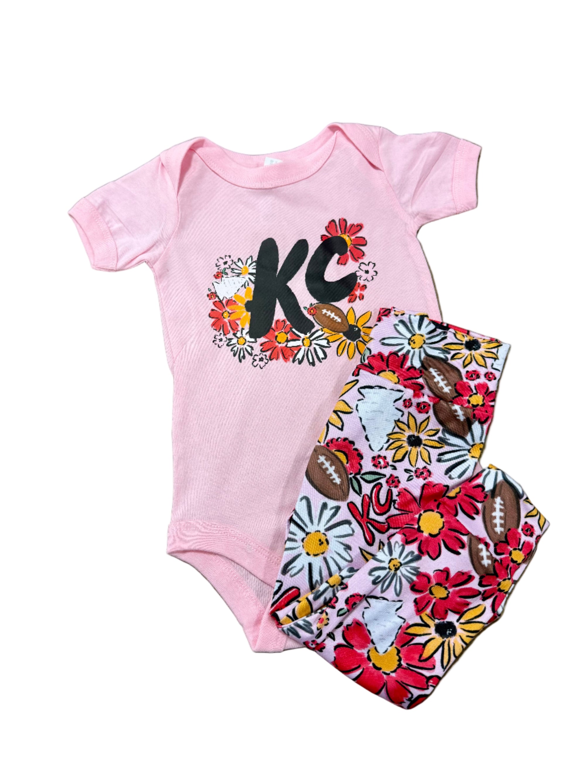 Pink baby snap bodysuit with 'KC' design and floral pants on a textured surface