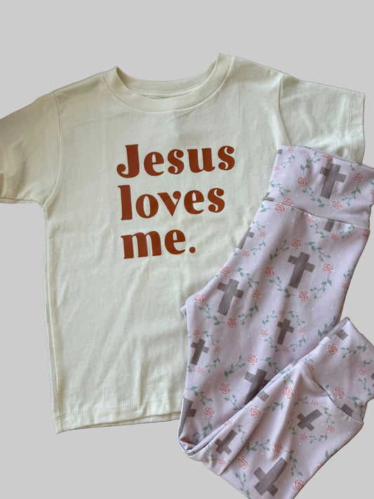 White top with 'Jesus loves me' text and a folded baby outfit with crosses on a wooden surface.
