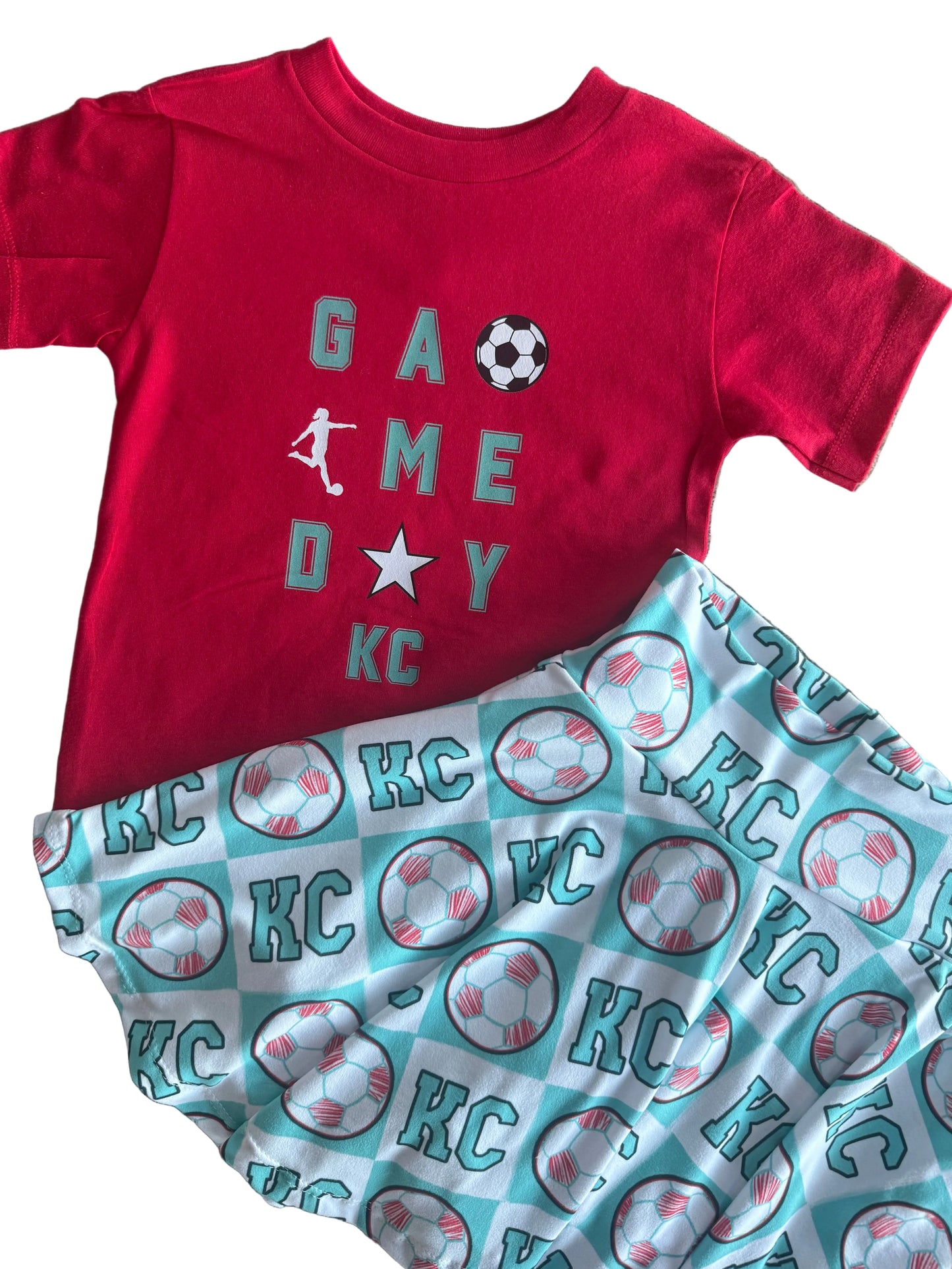 Red shirt with 'Game Day' and soccer sports-themed design, next to a patterned fabric with sports graphics on a textured surface.