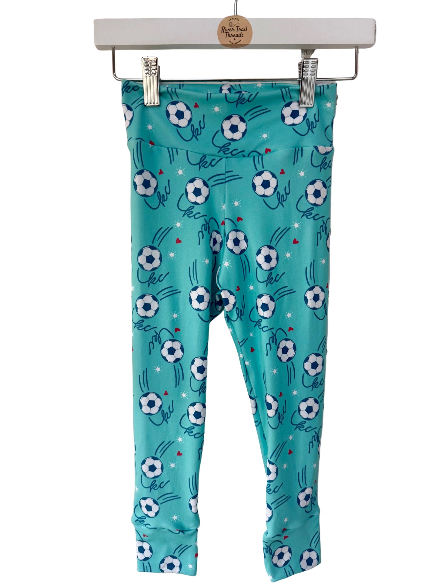 Teal KC leggings with soccer ball and heart pattern hanging on a brick wall.