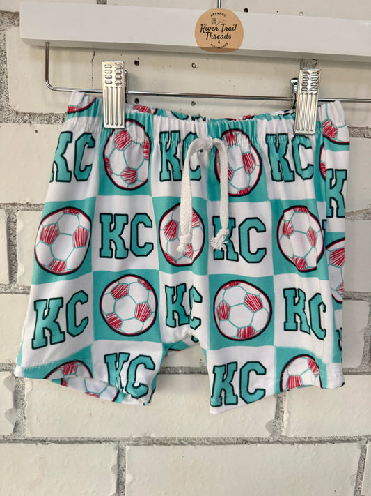 Pair of shorts with 'KC' and soccer ball pattern hanging on a rack.