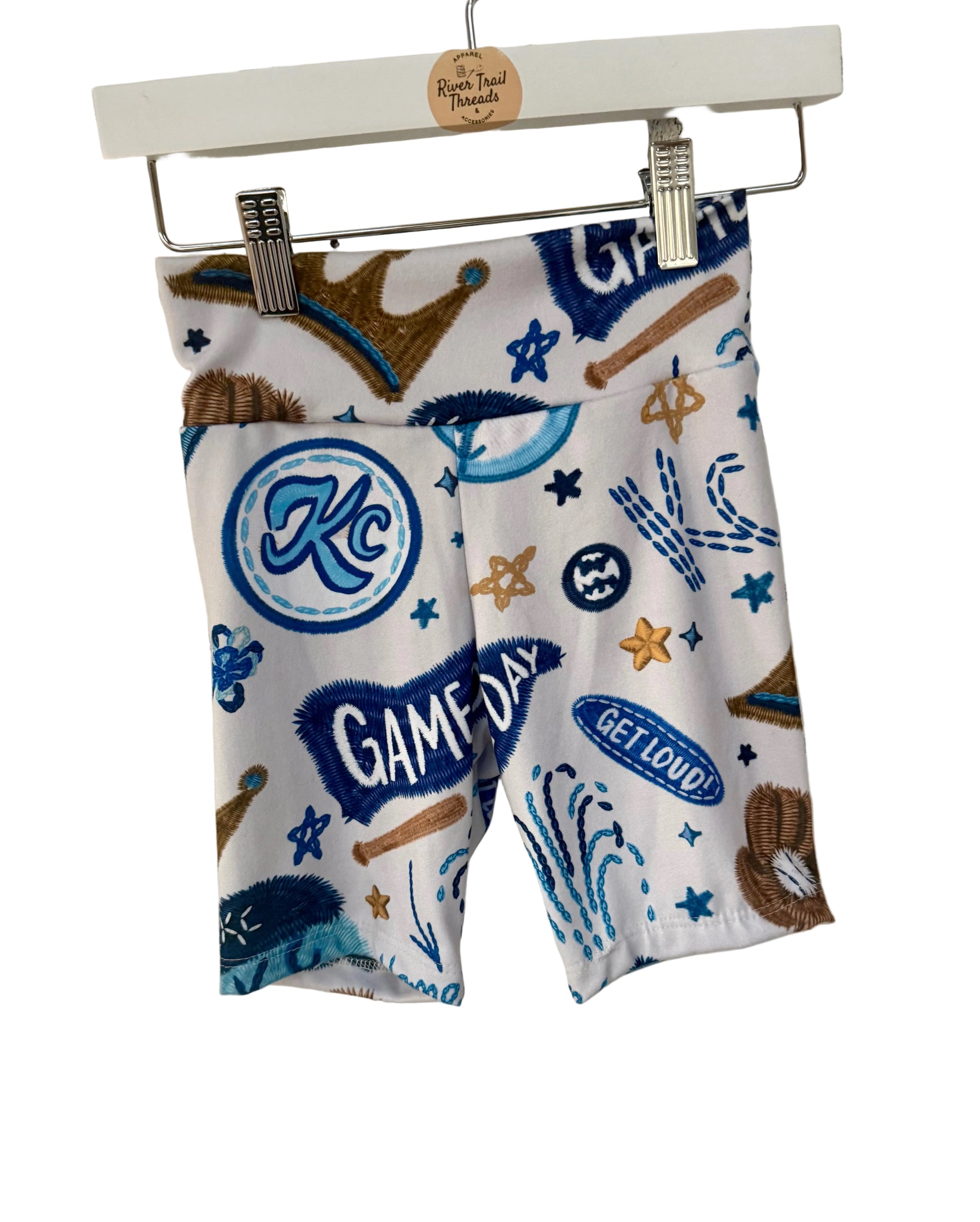 Pair of girls biker shorts with kc baseball sports-themed design hanging on a white brick wall.